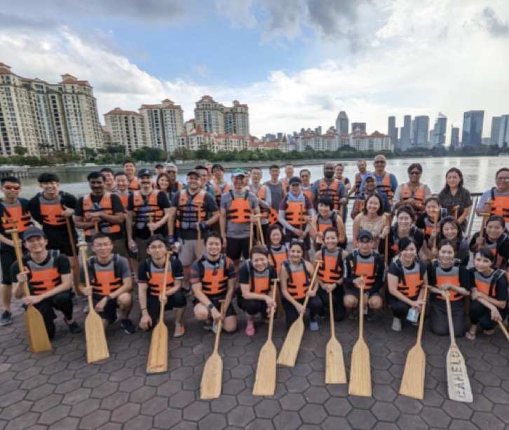 Gruppe von Mitarbeitenden in Schwimmwesten mit Bootspaddel vor einem See